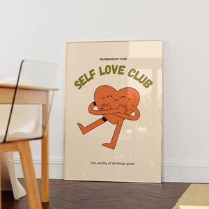 May include: A beige poster with a gold frame features a cartoon heart character with arms and legs hugging itself. The text "MEMBERSHIP PASS SELF LOVE CLUB" is in green letters above the heart. The text "I am worthy of all things good" is below the heart.
