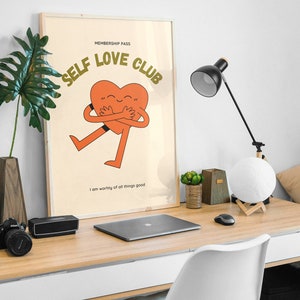 May include: A framed poster with a light brown background and the text "MEMBERSHIP PASS SELF LOVE CLUB" in green letters. A cartoon orange heart with a smiling face is hugging itself and has the text "I am worthy of all things good" below it.