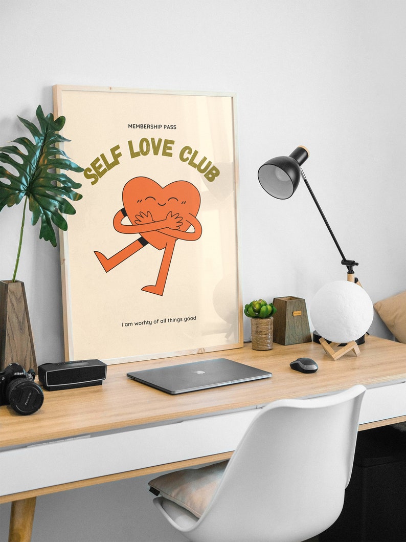 May include: A framed poster with a light brown background and the text "MEMBERSHIP PASS SELF LOVE CLUB" in green letters. A cartoon orange heart with a smiling face is hugging itself and has the text "I am worthy of all things good" below it.