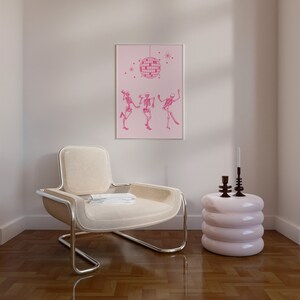 Puede incluir: Un sill&oacute;n blanco con marco cromado se encuentra en una habitaci&oacute;n con suelo de madera clara. Una mesa auxiliar rosa de tres niveles se encuentra junto al sill&oacute;n. Una impresi&oacute;n enmarcada con fondo rosa y una bola de discoteca colgando del techo con tres esqueletos bailando debajo est&aacute; colgada en la pared.