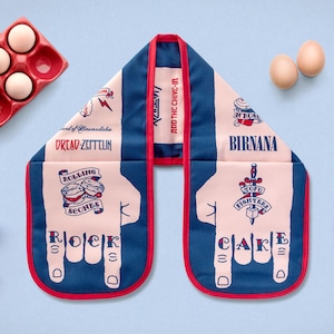 May include: Blue and pink oven mitt with a rock and roll theme. The mitt features hand illustrations spelling "ROCK CAKE" and band references. Includes eggs, flour, and an egg carton.