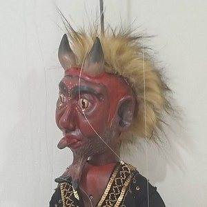 May include: A handcrafted devil puppet with a red face, black horns, and a faux fur hairstyle. The puppet wears a black garment with gold embellishments. The puppet is suspended by strings, suggesting it is a marionette.