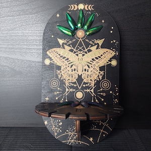 Puede incluir: Una estantería de madera negra con un diseño celestial. La estantería presenta una ilustración de polilla dorada, formas geométricas y patrones de estrellas. También hay alas de escarabajo verdes y una pequeña estantería. La estantería mide unos 30 cm de alto.
