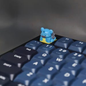 May include: A blue and yellow figurine of a cartoon character sits atop a mechanical keyboard. The keyboard has dark blue and navy keys. The figurine is centered on the keyboard, with the keys in focus. The background is a solid gray.