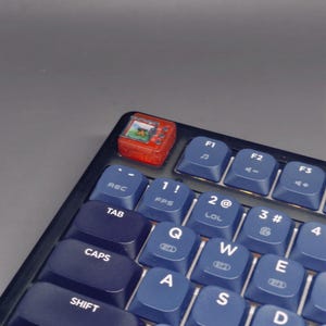 May include: A close-up of a black and blue mechanical keyboard with white lettering. A small red handheld gaming device sits on the top left corner. The keyboard keys include letters, numbers, and function keys.