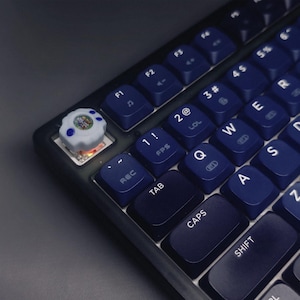 May include: Close-up of a mechanical keyboard with dark blue keycaps. A custom keycap with a white and blue design sits on the top left. The keyboard has a black frame and various function keys.