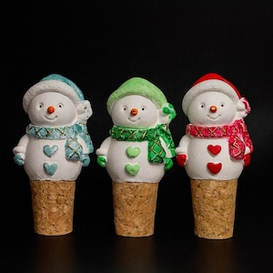 Könnte beinhalten: Drei Schneemann-Flaschenverschlüsse mit Korkböden. Jeder Schneemann hat einen andersfarbigen Hut und Schal: blau, grün und rot. Die Schneemänner haben herzförmige Knöpfe und Karottennasen. Der Hintergrund ist schwarz.