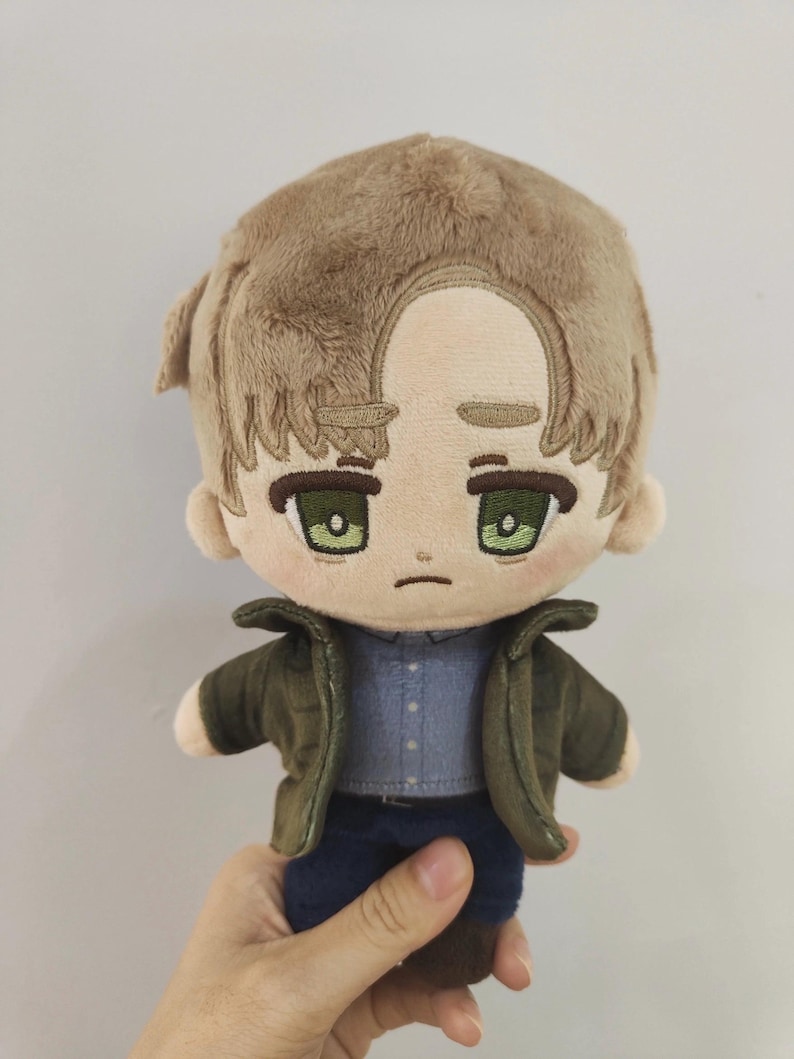 Op de afbeelding: Een pluche speelgoed van een cartoonfiguur met bruin haar, groene ogen en een neutrale uitdrukking. Het personage draagt een blauw shirt, een groen jack en een blauwe jeans.