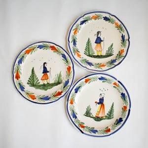 May include: Three white ceramic plates with blue, green, and orange painted borders. Each plate features a different hand-painted illustration of a person in a landscape setting.