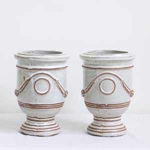 May include: Two matching off-white ceramic vases. Each vase has a cylindrical body with a decorative brown rope and circle design. The base and rim feature horizontal brown stripes. The vases are on a white surface.
