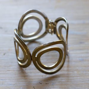 May include: A gold-toned metal cuff bracelet featuring a series of interconnected, open-work circular and oval shapes. The bracelet has a hinged opening mechanism and a polished finish that reflects light across its curved, organic-looking structure.