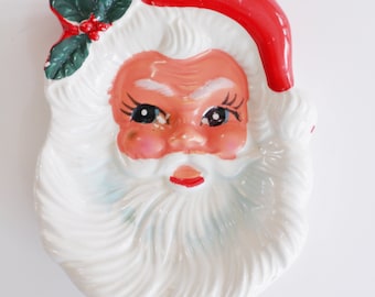 May include: A white ceramic Santa Claus dish with a red hat, white beard, and a green holly sprig.