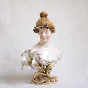 May include: A porcelain bust of a woman with a flower in her hair and a lily in her hand. The bust is painted in shades of white, pink, and green.