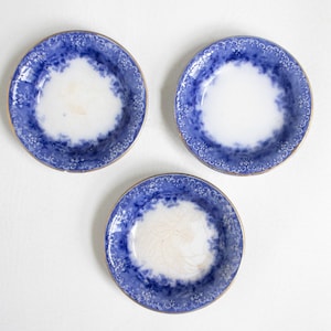 May include: Three round ceramic saucers featuring a white center and a wide, dark blue border decorated with a repeating floral pattern. Each saucer has a thin gold-toned rim. The bottom saucer displays visible fine crackling in the glaze.