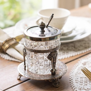 May include: A clear glass jam jar with a diamond-cut pattern, held in an ornate silver-toned metal frame with a matching circular base, lid, and small spoon. The metal components feature intricate floral engravings and a dark, aged patina finish.