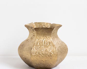 May include: A gold-colored metal vase with a wide, scalloped rim and a rounded body. The surface features an intricate embossed pattern of grape clusters and leaves, with a textured finish that covers the entire exterior of the vessel.