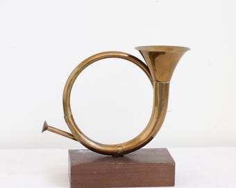May include: A polished brass hunting horn shaped object mounted on a rectangular dark brown wooden base. The brass instrument features a circular curved body with a wide flared bell opening at the top and a small pointed mouthpiece at the side.