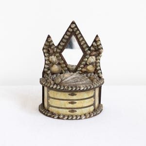 May include: A decorative jewelry box with a mirror, adorned with seashells. The box features three small drawers and a diamond-shaped mirror. The box is embellished with various seashells and has a light-colored, possibly pearlescent, finish.