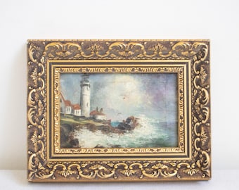 May include: A rectangular painting of a lighthouse on a rocky coastline with crashing waves, enclosed in an ornate, dark brown and gold-toned frame featuring intricate embossed scrollwork and decorative patterns along the border edges.