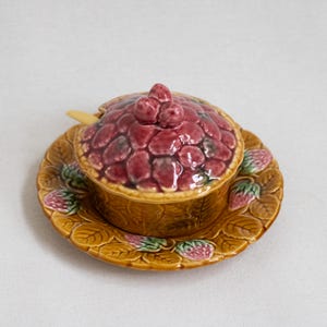 May include: A ceramic strawberry-themed serving dish with a lid, spoon, and matching saucer. The dish and saucer are brown with strawberry and leaf details. The lid is covered in red berry shapes.