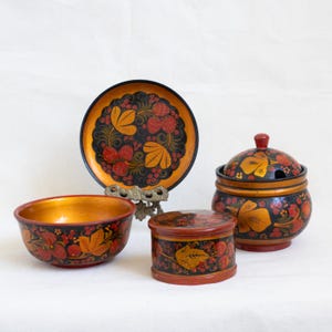 May include: A set of four hand-painted wooden bowls with a traditional Russian folk art design. The bowls are decorated with red, orange, and black floral patterns and feature a gold background. The set includes a large plate, a small bowl, a sugar bowl with a lid, and a small round box.