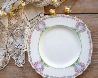 May include: A white ceramic plate with a scalloped edge, featuring a decorative border of pink roses and green foliage. The rim is accented with small yellow floral details and a thin green line, surrounding a plain white center.