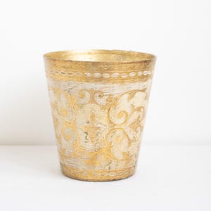 May include: A decorative, cylindrical container with a gold rim and base. The container features a cream-colored background with ornate gold floral scrollwork. The design is reminiscent of antique or vintage decor.