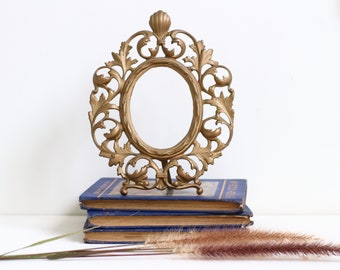 May include: A gold-toned metal picture frame with an ornate design. The frame is oval-shaped and has a stand on the bottom. The frame is sitting on top of a stack of blue books.