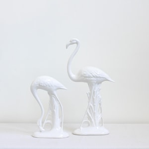 May include: Two white ceramic flamingo figurines standing on stylized bases. One flamingo is depicted with its neck arched downward, while the other stands tall with its neck curved upward. Both figures feature detailed feather textures and smooth finishes.