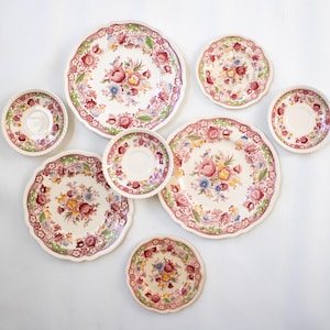 May include: A collection of vintage floral patterned plates and saucers. The plates and saucers feature a cream background with a red, pink, yellow, and blue floral design. The edges of the plates have a scalloped design.