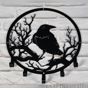 May include: Black metal wall-mounted key holder shaped like a circle with a raven perched on a branch. The raven is silhouetted against a white brick wall.