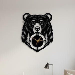 Puede incluir: Reloj de pared con cabeza de oso negro con una esfera con manecillas doradas y fondo blanco. El reloj está cortado de una sola pieza de material y está montado en una pared blanca. El reloj es un artículo decorativo.