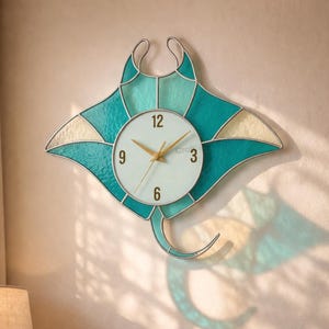 May include: A wall clock shaped like a manta ray, featuring a teal and cream stained glass design. The clock face is white with gold hands and black numerals. The clock is mounted on a beige wall above a wooden bed frame.