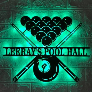 May include: A black metal sign with a green light behind it. The sign features a pool table with crossed cues and the text "Leeray's Pool Hall".
