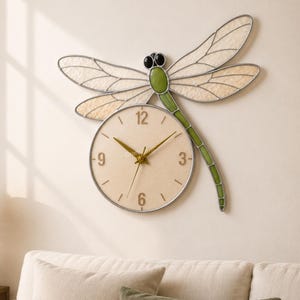 Könnte beinhalten: Wanduhr in Libellenform mit cremefarbenem Zifferblatt und goldenen Zeigern. Die Libelle hat grüne und cremefarbene Flügel und einen Körper. Die Uhr ist an einer beigefarbenen Wand über einem Sofa befestigt.