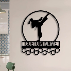 Może przedstawiać: Czarna metalowa grafika ścienna przedstawiająca postać karate w wysokim kopnięciu w okręgu. Poniżej figury znajduje się prostokątne miejsce na niestandardową nazwę, z kilkoma okrągłymi uchwytami poniżej. Grafika jest zamontowana na jasnej ścianie.