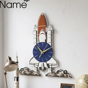 May include: A wooden wall clock shaped like a space shuttle, with a blue clock face and gold hands. The clock is white, orange, and black. The clock is mounted on a white wall. The word "CraftName" is above the clock.