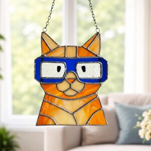 May include: Stained glass cat suncatcher with orange and yellow hues, wearing blue glasses. The cat is suspended by a chain. The sign at the bottom says "CraftName sign".
