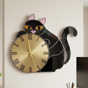 Könnte beinhalten: Eine schwarze Wanduhr in Katzenform mit goldfarbenem Zifferblatt und römischen Ziffern. Die Ohren der Katze sind rosa und ihre Augen gelb. Die Uhr ist an einer weißen Wand über einem Holzschreibtisch angebracht. Die Uhr hat einen Durchmesser von ca. 30 cm.