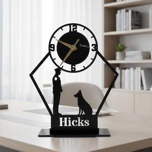 May include: Black desk clock featuring a police officer and dog silhouette. The clock has a circular face with gold hands and a black frame. The name "Hicks" is displayed below the figures. The clock is on a wooden desk.