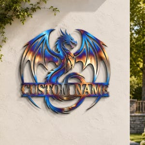 May include: A metal dragon wall sign with the text "CUSTOM NAME". The dragon is in shades of blue, gold, and bronze. The sign is mounted on a white wall, with greenery and flowers in the background.