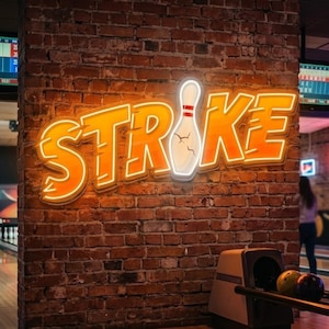 May include: An orange neon sign spelling "STRIKE" with a white bowling pin graphic, mounted on a brick wall. The sign is in a bowling alley. The sign is a product of CraftName sign.