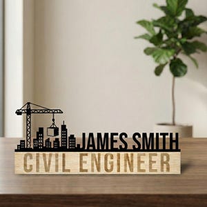 Può includere: Un cartello da scrivania in legno con il nome "JAMES SMITH" e il titolo "CIVIL ENGINEER" incisi in legno chiaro. Una silhouette nera di un paesaggio urbano con una gru è sopra il nome. Il cartello è su una superficie di legno.