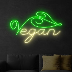 May include: A neon sign with the word "vegan" in a cursive font. The word is outlined in yellow and filled in with green. The sign is mounted on a black wall above a black couch.