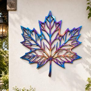 May include: A large, metal maple leaf wall art piece with a vibrant colour gradient of blue, purple, and gold. The leaf is mounted on a white wall, with the text "CraftName" visible at the bottom. The art piece is an outdoor decoration.