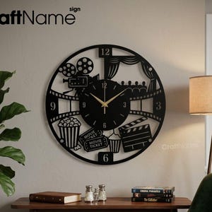 May include: Black metal wall clock with a movie theme. The clock features a film reel, popcorn, movie ticket, and a clapperboard. The clock hands are gold. The text "CraftName" is visible in the upper left and lower right corners.