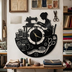 May include: Black metal wall clock featuring a silhouette of a seamstress at a sewing machine. The clock face includes numbers, a ruler, and spools of thread. The clock is surrounded by sewing tools and is mounted on a white wall.