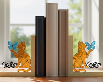 Puede incluir: Un par de sujetalibros de madera con un diseño de gato naranja sosteniendo una mariposa azul. El gato tiene un patrón geométrico. Los sujetalibros tienen el texto "Custom name" y "CraftName sign". Libros colocados entre los sujetalibros.