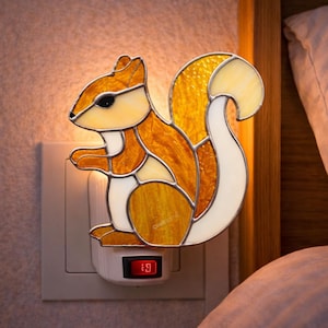 Puede incluir: Una luz de noche de vidriera con forma de ardilla, con tonos naranja, amarillo y blanco. La ardilla está enchufada a una toma de corriente con un interruptor rojo iluminado. La marca "CraftName" se muestra en la parte inferior.