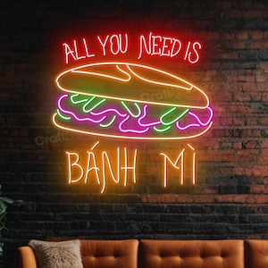 May include: A neon sign on a brick wall reads "ALL YOU NEED IS" above a colorful illustration of a Bánh mì sandwich, with the word "BÁNH MÌ" below. The sign is above a brown sofa with a fluffy pillow.